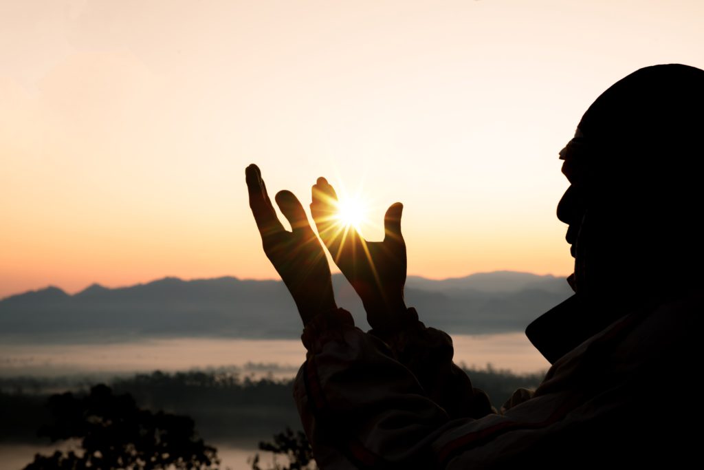 Em local alto e com paisagem de pôr do sol, silhueta de homem ergue mãos ao céu - bênçãos