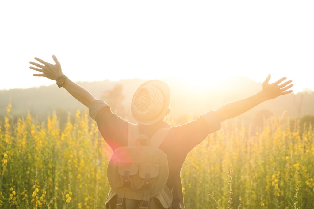 Homem que leva mochila nas costas abre os braços em direção ao sol em campo com vegetação alta