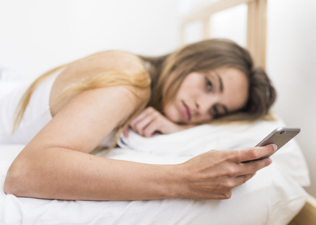 Mulher loira segura celular deitada na cama e com aparente preocupação