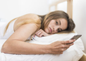 Mulher loira segura celular deitada na cama e com aparente preocupação