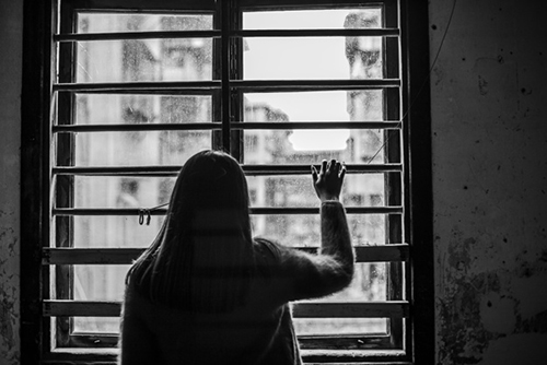 Foto em preto e branco de mulher que olha para fora através de vidros de uma janela grande